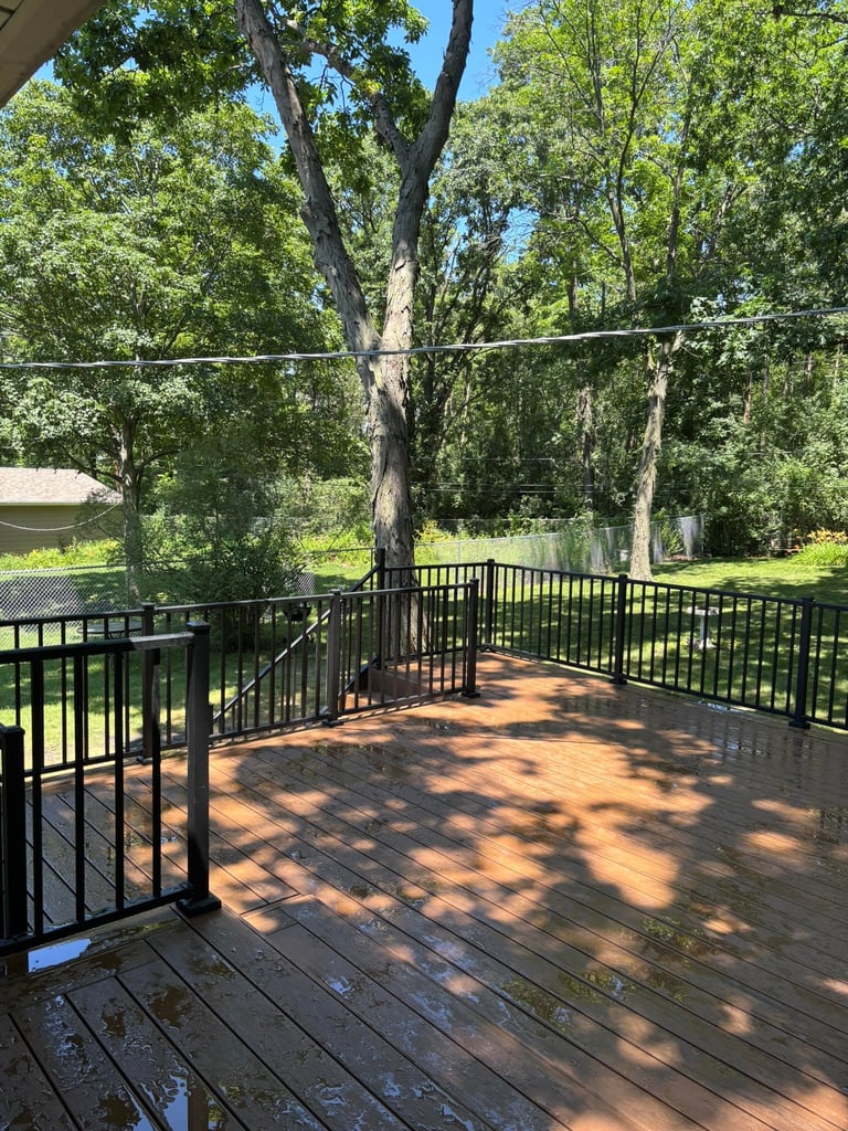 Wooden deck with black metal railing overlooking lush green trees and backyard on sunny day