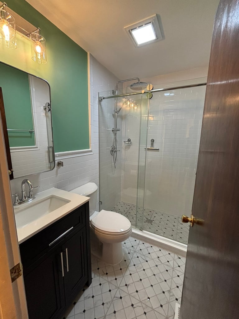 Modern bathroom with green and white walls, black vanity, toilet, glass shower enclosure, and patterned tile floor.
