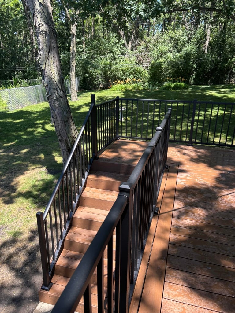 Wooden deck with black metal railings surrounded by lush green lawn and tall trees with dappled sunlight