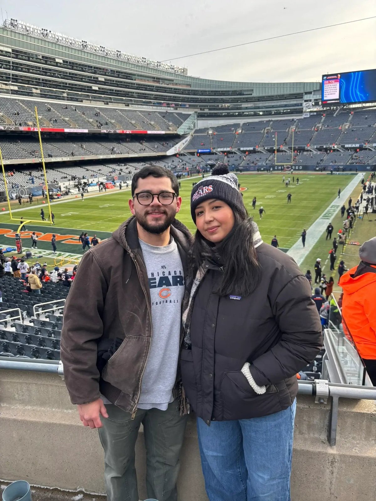 Two people posing at a large football stadium during a game, with the field and crowd visible in the background