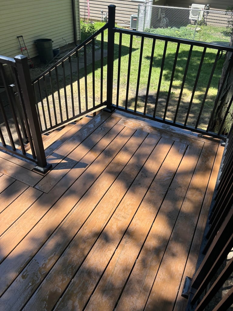 Black railed deck with composite wood-colored boards and shadow patterns, attached to a green-sided house with fenced yard background