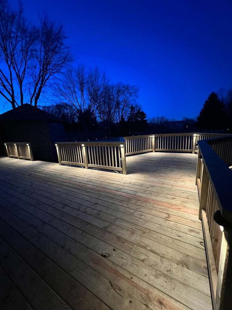 Illuminated wooden deck at dusk with white railings overlooking bare trees and dark blue sky at twilight