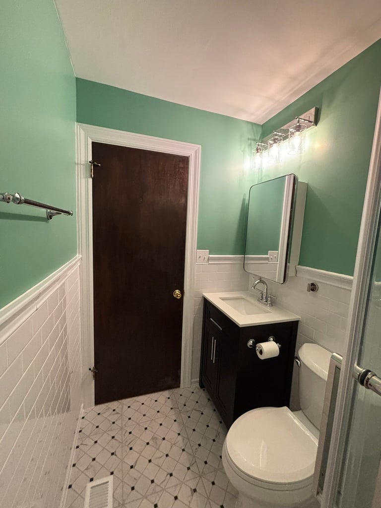 Modern bathroom with mint green upper walls, white subway tile wainscoting, black door, vanity with sink, mirror, toilet, and checkered floor