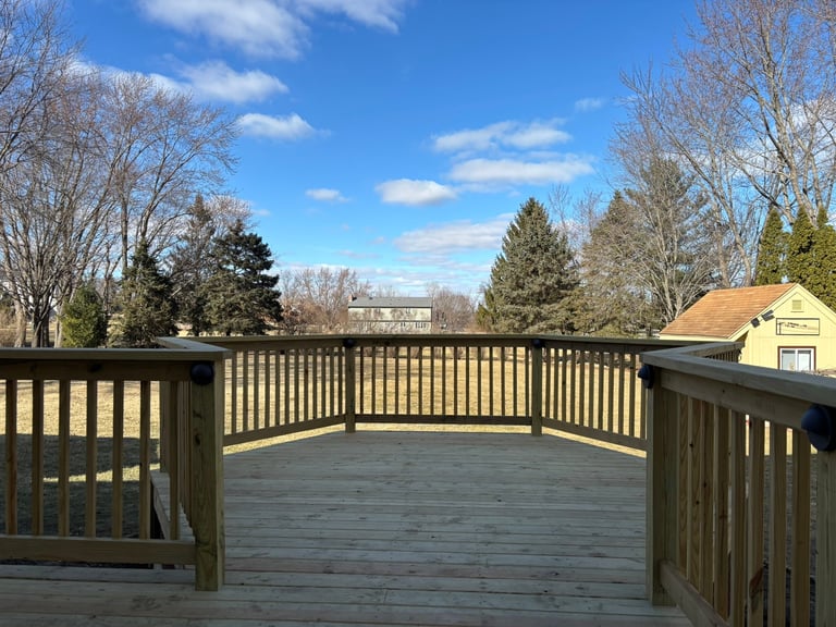 Large wooden deck with dark railings overlooking bare trees and residential houses under a clear blue sky