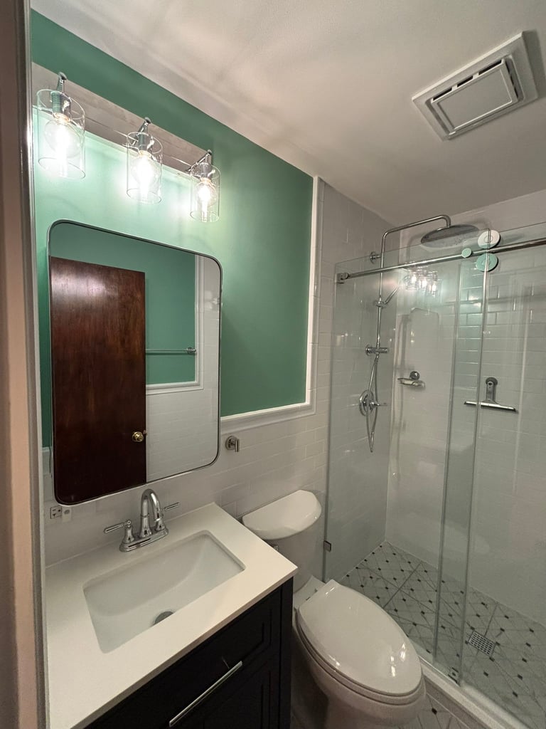 Modern bathroom with green accent wall, white sink vanity, illuminated mirror, toilet, and glass walk-in shower enclosure