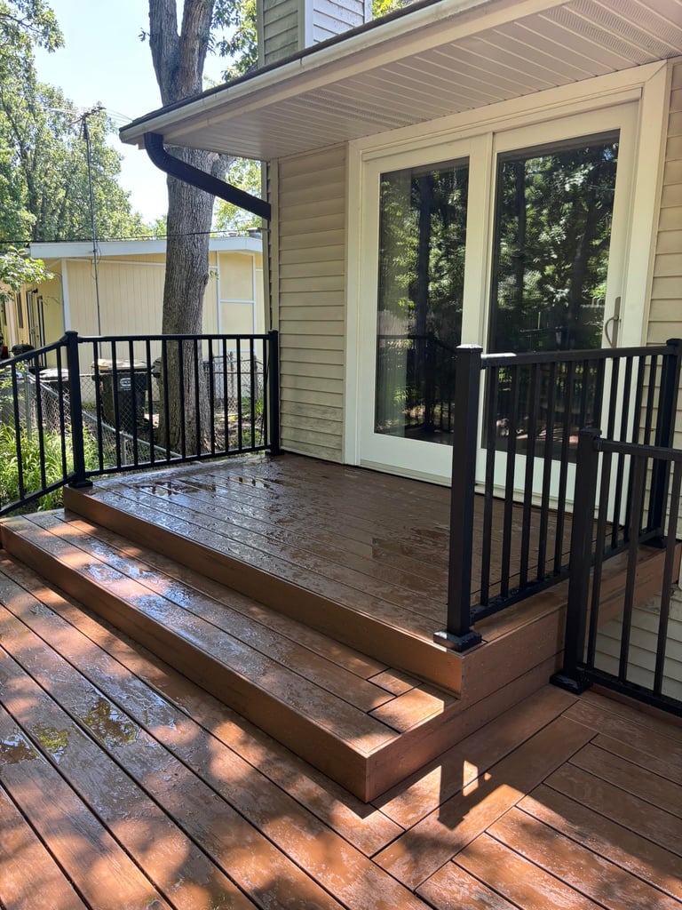Modern wooden deck with black railings attached to beige house with large windows and pergola, surrounded by trees